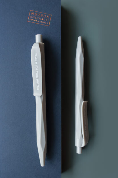 Dwa szare długopisy o lekko geometrycznych kształtach. Na klipsie długopisu połyskujący napis Muzeum Emigracji w Gdyni.