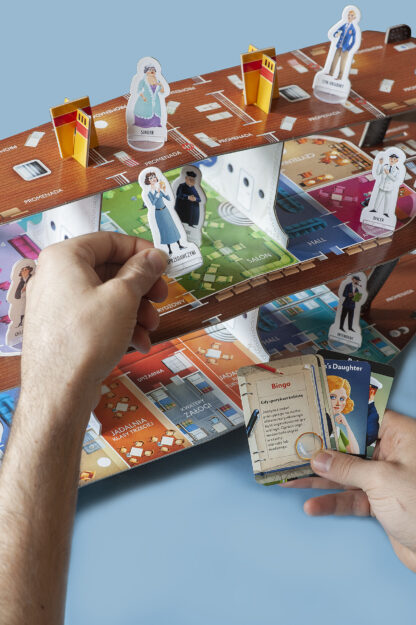 Mężczyzna w jednej dłoni trzyma karty do gry. W drugiej dłoni trzyma pionka – kartonowy wizerunek jednej z postaci.