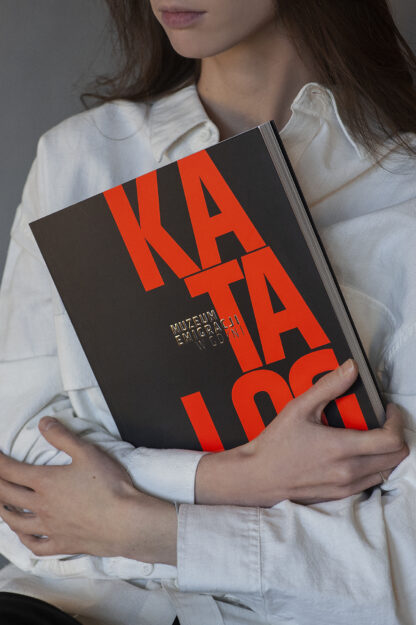Kobieta ubrana w białą koszulę trzyma dużą książkę. Na czarnej okładce pomarańczowy napis: Katalog. Muzeum Emigracji Gdyni.