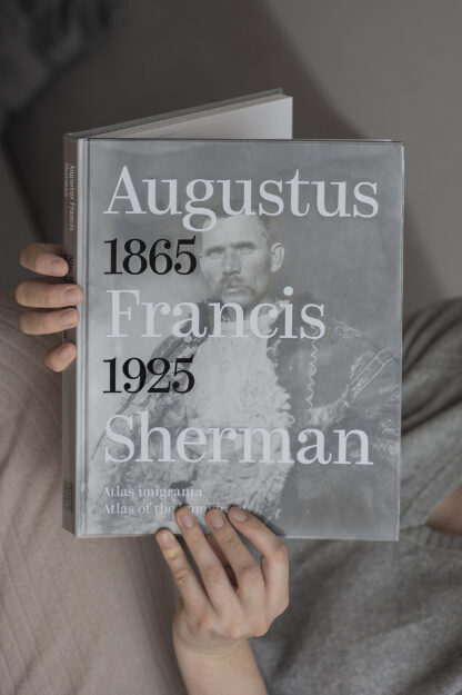 Otwarta książka trzymana w dłoniach. Na okładce napis Augustus Francis Sherman, 1865 – 1925, Atlas imigranta. W tle jedno z czarno-białych zdjęć Shermana. Na zdjęciu rumuński pasterz – mężczyzna z wąsem, ubrany w gruby płaszcz i biały sweter.