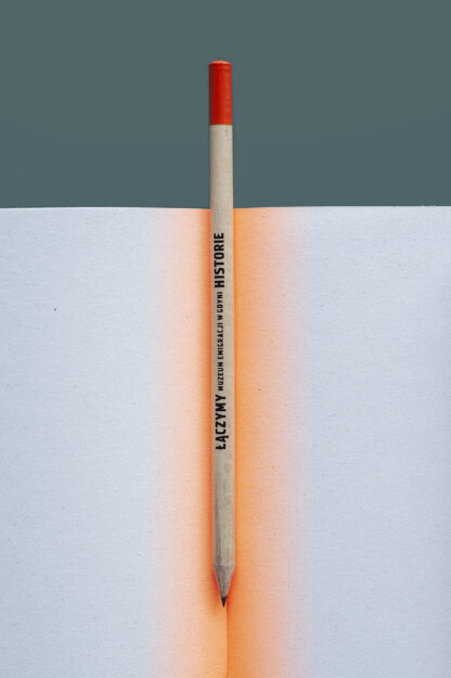 Ołówek umieszczony w środku otwartego notatnika. Końcówka ołówka wystaje u góry notatnika. Ołówek ma czarny napis Muzeum Emigracji w Gdyni, łączymy historie. Koniec ołówka w jaskrawym, pomarańczowym kolorze.