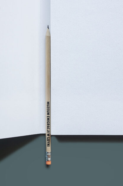 Ołówek umieszczony w środku otwartego notatnika. Dolna część wystaje poza notatnik. Ołówek drewniany z czarnym napisem: Muzeum Emigracji w Gdyni. Na końcu ołówka gumka w jaskrawym, pomarańczowym kolorze.