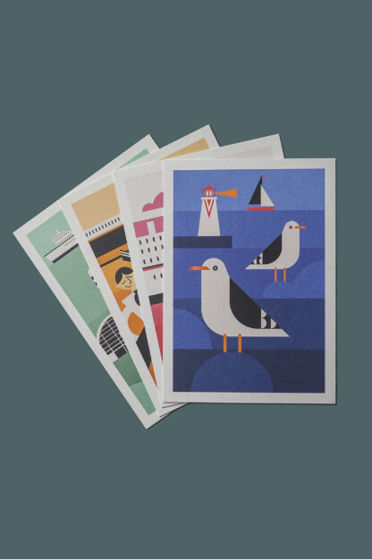 Cztery pocztówki z grafikami w różnych kolorach, ułożone jedna na drugiej. Na wierzchu niebieska pocztówka. Na niej dwie mewy, w oddali latarnia morska oraz żaglówka.