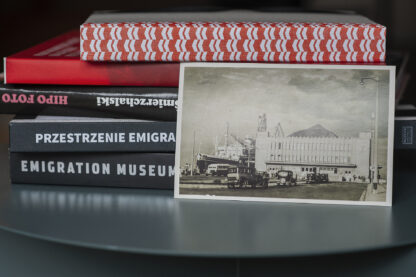 Pocztówka oparta o książki. Przedwojenne czarno-białe zdjęcie Dworca Morskiego w Gdyni. Przed budynkiem samochody, a po jego lewej stronie statek pasażerski.