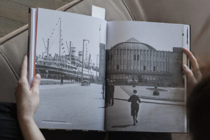 Osoba trzyma w rękach otwartą książkę. Obie strony zajmuje czarno-białe zdjęcie. Na zdjęciu budynek Dworca Morskiego z napisem Gdynia Ameryka na oknach. Po lewej stronie statek pasażerski stojący przy nabrzeżu. Przy statku tłum ludzi. Na placu przed budynkiem pojedyncze osoby.