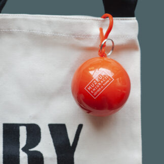 Pomarańczowy pojemnik w kształcie kuli z logo Muzeum Emigracji. Pojemnik ma uchwyt zaczepiony o kremową torbę.