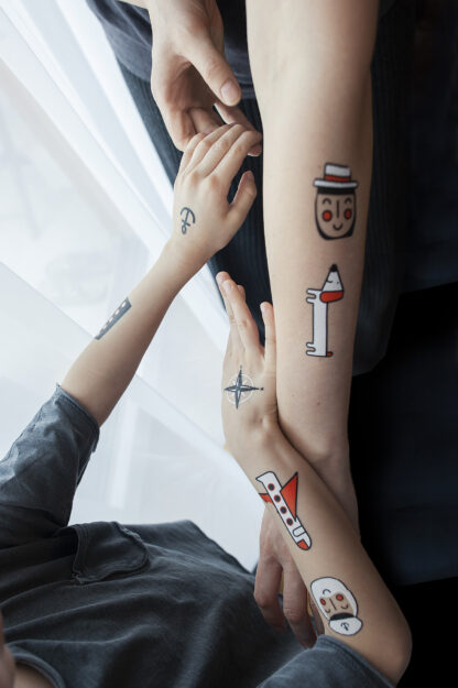 Dwie osoby – dziecko i osoba dorosła. Mają wyciągnięte ku sobie ręce, stykają się dłońmi. Na rękach mają zmywalne tatuaże o różnych kształtach.