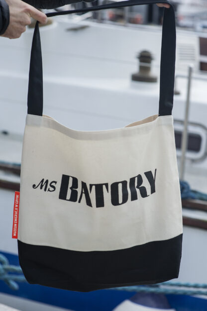 Mężczyzna trzyma czarno-białą torbę z czarnym paskiem na ramię. Na środku torby napis MS Batory. W tle gdyńska marina.