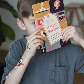 Chłopiec trzyma kolorowy zeszyt, zasłaniając sobie większość twarzy. Na środku zeszytu narysowana zawieszka z napisem wyglądającym na odręczny: Zeszyt podróżny. Wokół zawieszki kolorowe kwadraty z grafikami o tematyce podróżniczej.