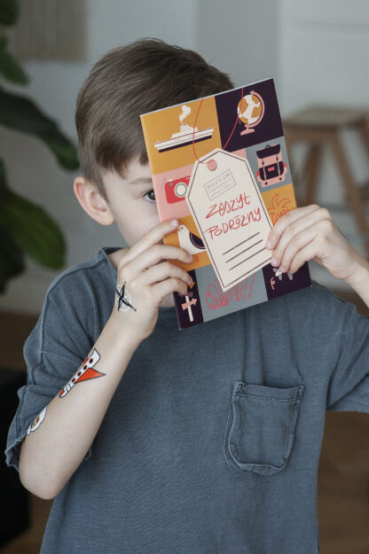 Chłopiec trzyma kolorowy zeszyt, zasłaniając sobie większość twarzy. Na środku zeszytu narysowana zawieszka z napisem wyglądającym na odręczny: Zeszyt podróżny. Wokół zawieszki kolorowe kwadraty z grafikami o tematyce podróżniczej.