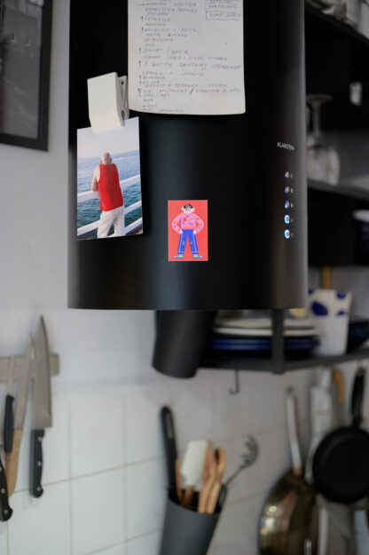 Magnes z ilustracją wisi na okapie w kuchni. Na ilustracji na czerwonym tle Franek – bohater wystawy. Ubrany w różowy sweter i niebieskie spodnie. Stoi z założonymi na biodra rękami.